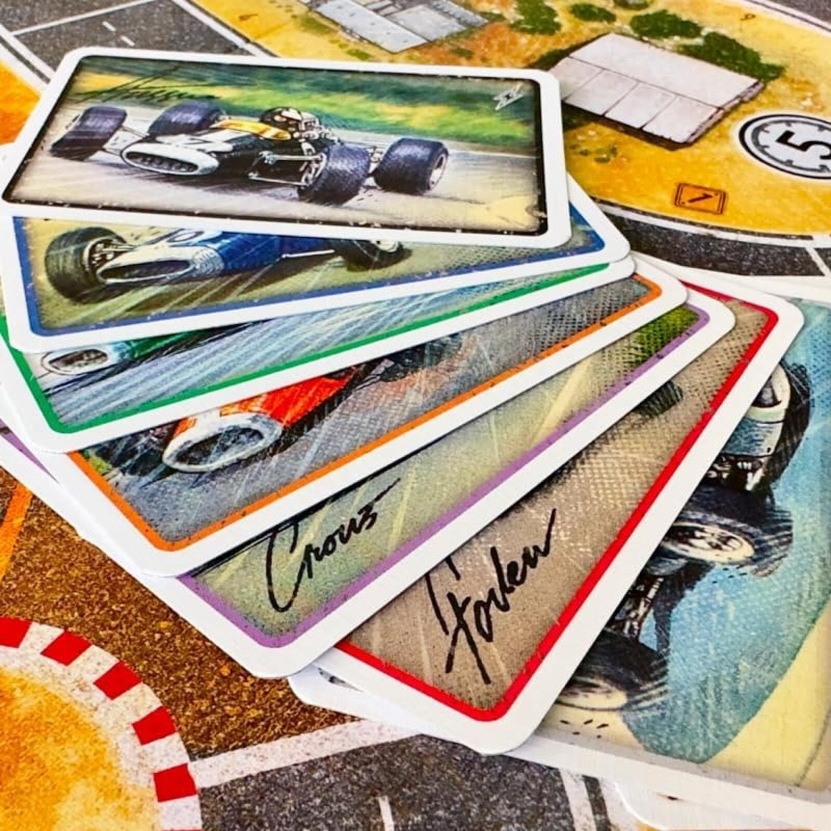 A hand holding two cards from the board game Catan, showing resource and action cards over a green and gray game board.