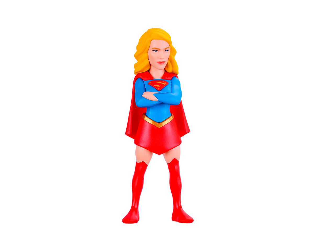 A small plastic figurine of a female superhero with blonde hair, wearing a blue and red costume with a red cape and boots, standing with arms crossed on a plain white background.